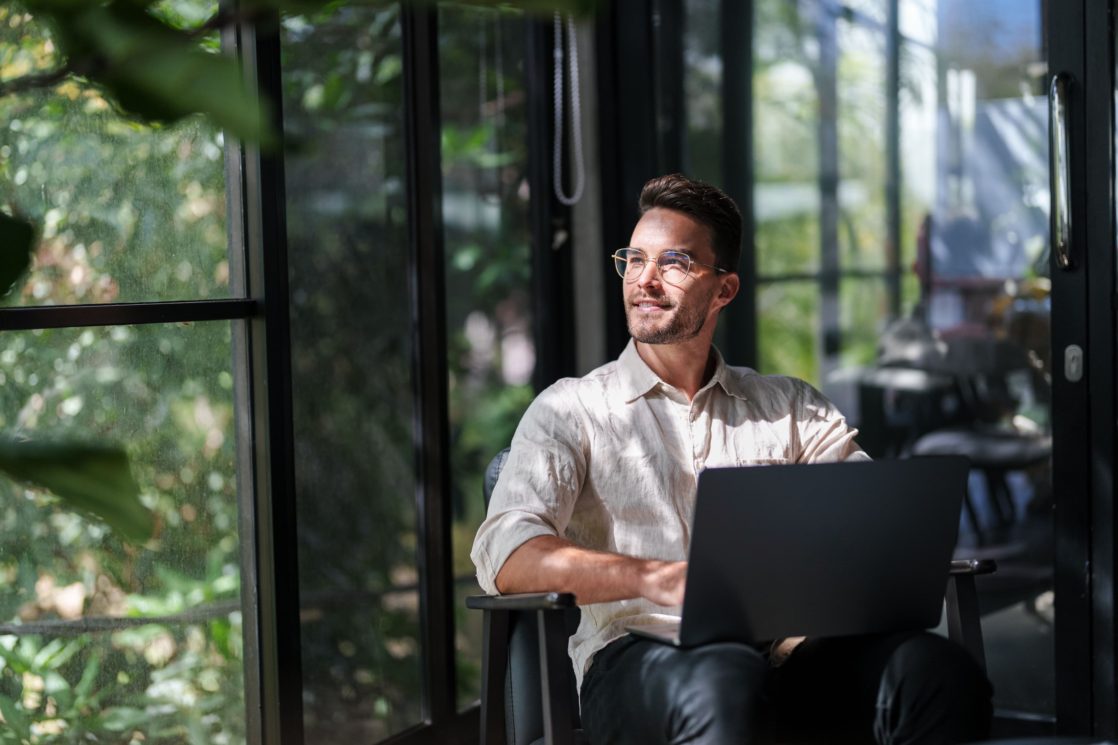 A professional pauses thoughtfully with a laptop in a bright, plant-filled space