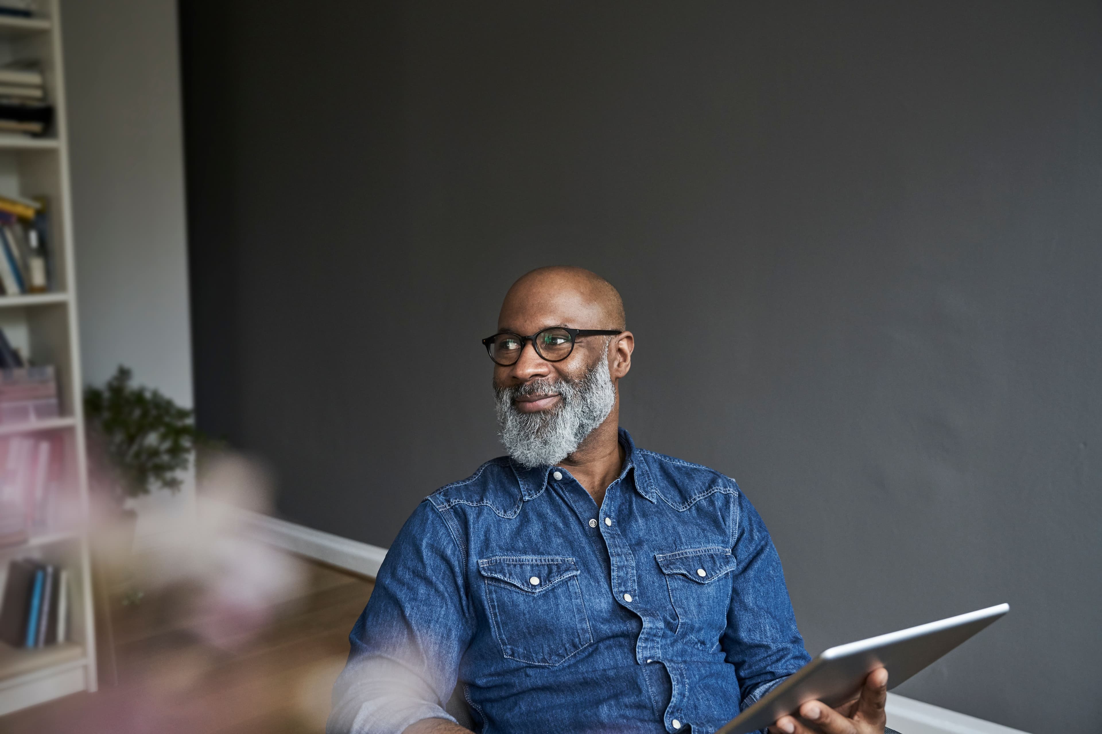 A man wearing glasses holds a tablet while looking toward someone nearby.